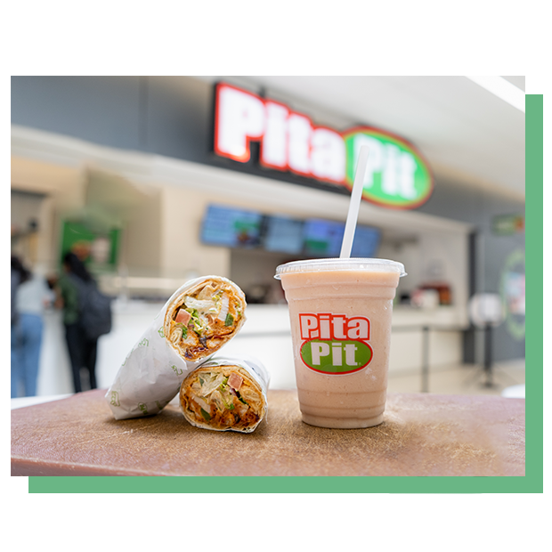 Two wraps and a shake on the table with pita pit store in the back